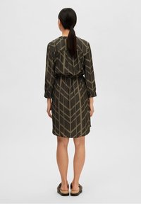 Vista posteriore di un vestito con motivo caratterizzato da un design geometrico verde e nero, maniche lunghe e vita elastica, abbinato a sandali piatti.