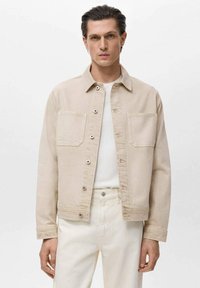 Beige spijkerjasje met een kraag, twee voorzakken en knoopsluitingen. Draagt over een wit shirt en gecombineerd met lichtgekleurde broeken.