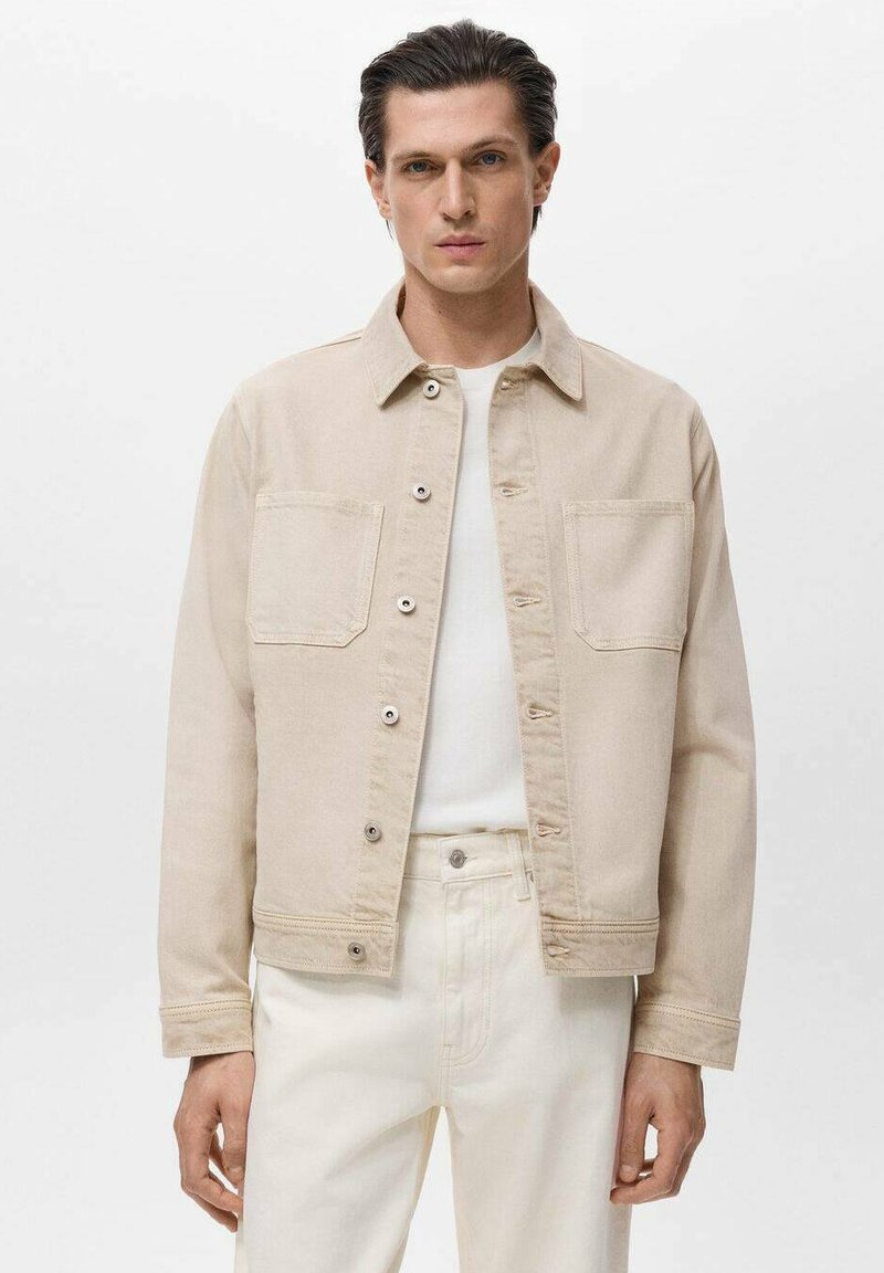 Beige spijkerjasje met een kraag, twee voorzakken en knoopsluitingen. Draagt over een wit shirt en gecombineerd met lichtgekleurde broeken.