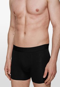 Bóxer de algodón negro con una suave cinturilla elástica que presenta una marca en relieve. Textura suave, diseño ajustado y longitud hasta medio muslo.