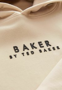Hoodie beige con una texture liscia, caratterizzato da un logo in rilievo nero che riporta la scritta "BAKER" e "by TED BAKER" sul davanti.