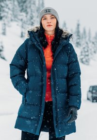 Tmavě modrý péřový kabát s kapucí lemovanou kožešinou, oranžová vnitřní vrstva a černé rukavice. Sněhové pozadí dodává scéně texturu.