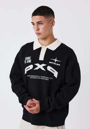 Jeune homme aux cheveux courts portant un pull noir avec un col blanc et le texte "PX9" et "Change The Game", regardant sur le côté, les mains jointes.