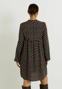 Robe noire avec un motif floral en losange, manches longues, dos plissé et coupe ample. Texture lisse, longueur genou, design minimaliste.