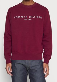 Bordowy sweatshirt z okrągłym dekoltem, ściągaczami przy rękawach i u dołu, z białym haftowanym logo "TOMMY HILFIGER" oraz datą założenia 1985.