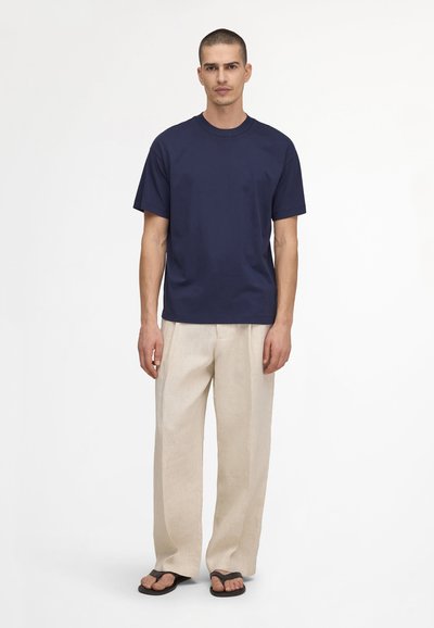 Man standing wearing a navy blue short-sleeve t-shirt, beige wide-leg pants, and black flip-flop sandals against a white background.
