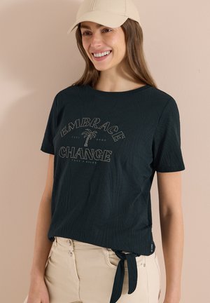Femme souriante portant une casquette beige, un t-shirt noir avec le texte « Embrace Change » et un motif de palmier, assorti à un pantalon beige sur un fond neutre.
