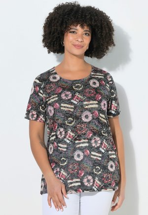 Kobieta o kręconych włosach, ubrana w czarny t-shirt z krótkim rękawem z kolorowymi abstrakcyjnymi okrągłymi i liniowymi wzorami, w połączeniu z białymi spodniami.