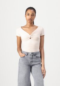 Top corti manica corta beige chiaro con un dettaglio torcibile sulla parte frontale, abbinato a jeans in denim grigio a vita alta e a gamba larga. Tessuto liscio, design aderente.