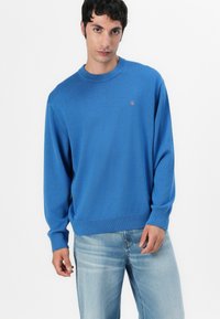 Maglione lavorato a maglia blu con polsini e bordo a coste, scollo a girocollo, caratterizzato da un piccolo logo ricamato sul petto. Indossato con jeans blu chiaro.