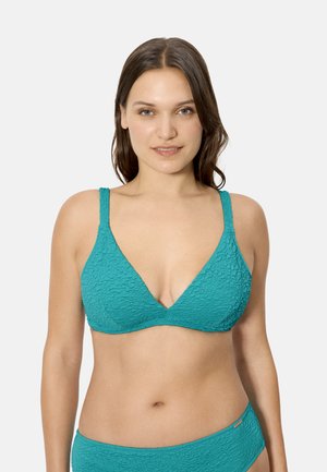 Mujer con cabello castaño hasta los hombros, vistiendo un top de bikini texturizado en color verde azulado y una parte inferior a juego, posando frente a un fondo blanco liso.