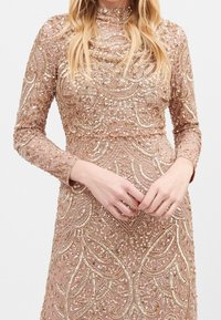 Perlesmykket aftenkjole i blød pink. Har detaljeret guldbroderi og pailletter, lange ærmer og en tætsiddende silhuet.