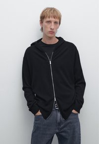 Schwarze Kapuzenshirtjacke aus weichem Stoff mit lockerer Passform. Verfügt über eine Kapuze, lange Ärmel und gerippte Bündchen. Wird über einem schwarzen T-Shirt getragen.