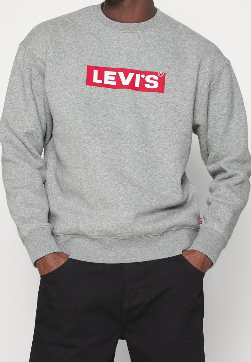 Homme portant un sweat-shirt gris avec un logo Levi's rouge sur la poitrine et un pantalon noir, les mains dans les poches.