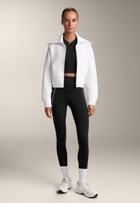 Giacca bianca con zip sopra un top nero corto, abbinata a leggings neri a vita alta con strisce laterali bianche. Scarpe sportive bianche con calze.