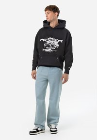 Zwarte hoodie met een grafisch ontwerp aan de voorkant en een buidelzak, gecombineerd met lichtblauwe jeans en zwart-witte sneakers.
