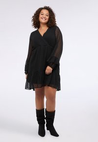 Vestido corto negro con diseño envolvente, patrón de puntos, mangas largas y dobladillo con volantes; combinado con botas negras caídas hasta la rodilla.