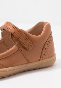 Zapato de cuero marrón claro con una correa de Velcro, acentos perforados y una suela de goma texturizada. Diseño minimalista con forma redondeada.