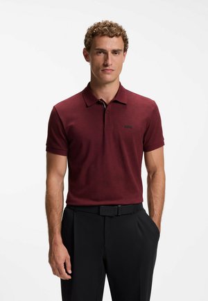 Mann mit lockigem Haar, der ein burgunderfarbenes kurzärmeliges Poloshirt und schwarze Hose mit schwarzem Gürtel vor einem einfarbigen weißen Hintergrund trägt.