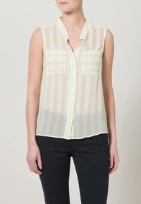 Blusa senza maniche di un delicato giallo chiaro con righe verticali, chiusura a bottoni e due tasche sul petto. Tessuto trasparente con una texture liscia.