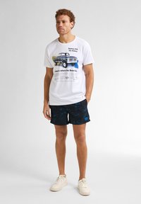 Biała koszulka z grafiką samochodu i napisem, zestawiona z czarnymi szortami kąpielowymi ozdobionymi niebieskimi wzorami kwiatowymi. Look dopełniają trampki.