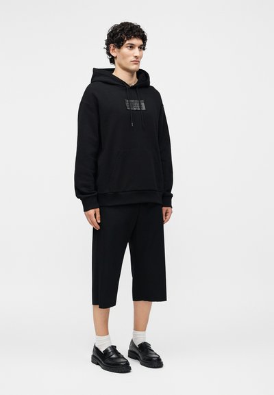 Ένας νεαρός άνδρας στέκεται facing μπροστά φορώντας μαύρη φόρμα με μπροστινή τσέπη, μαύρο ευρύ cropped παντελόνι, λευκές κάλτσες και μαύρα loafers.