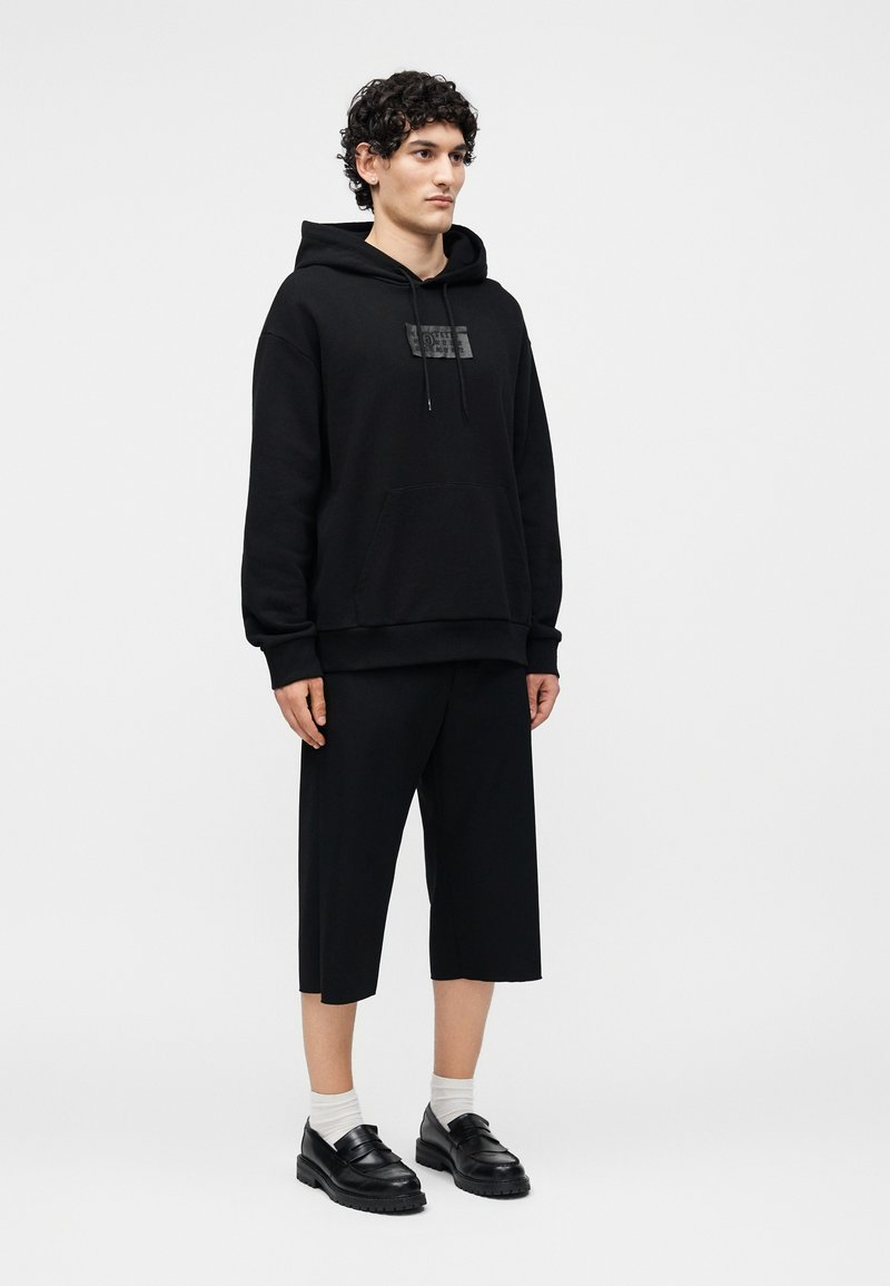 Ein junger Mann steht frontal, trägt einen schwarzen Hoodie mit Fronttasche, schwarze weite, gekürzte Hosen, weiße Socken und schwarze Loafers.