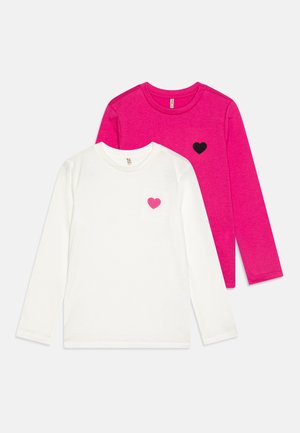 Deux t-shirts à manches longues : l'un blanc avec une poche en forme de cœur rose, l'autre rose avec un détail en forme de cœur noir. Les deux sont fabriqués en coton doux.