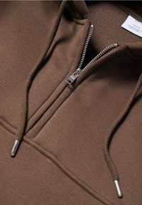 Felpa con zip di colore marrone realizzata in tessuto morbido; presenta un colletto a metà zip e lacci con dettagli metallici. Finitura liscia e texturizzata.