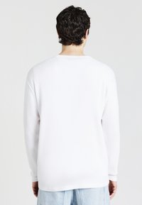 Camicia bianca a maniche lunghe con un design testurizzato e a coste. Collo rotondo, vestibilità rilassata e polsini sulle maniche. Indossata con jeans azzurri chiari.