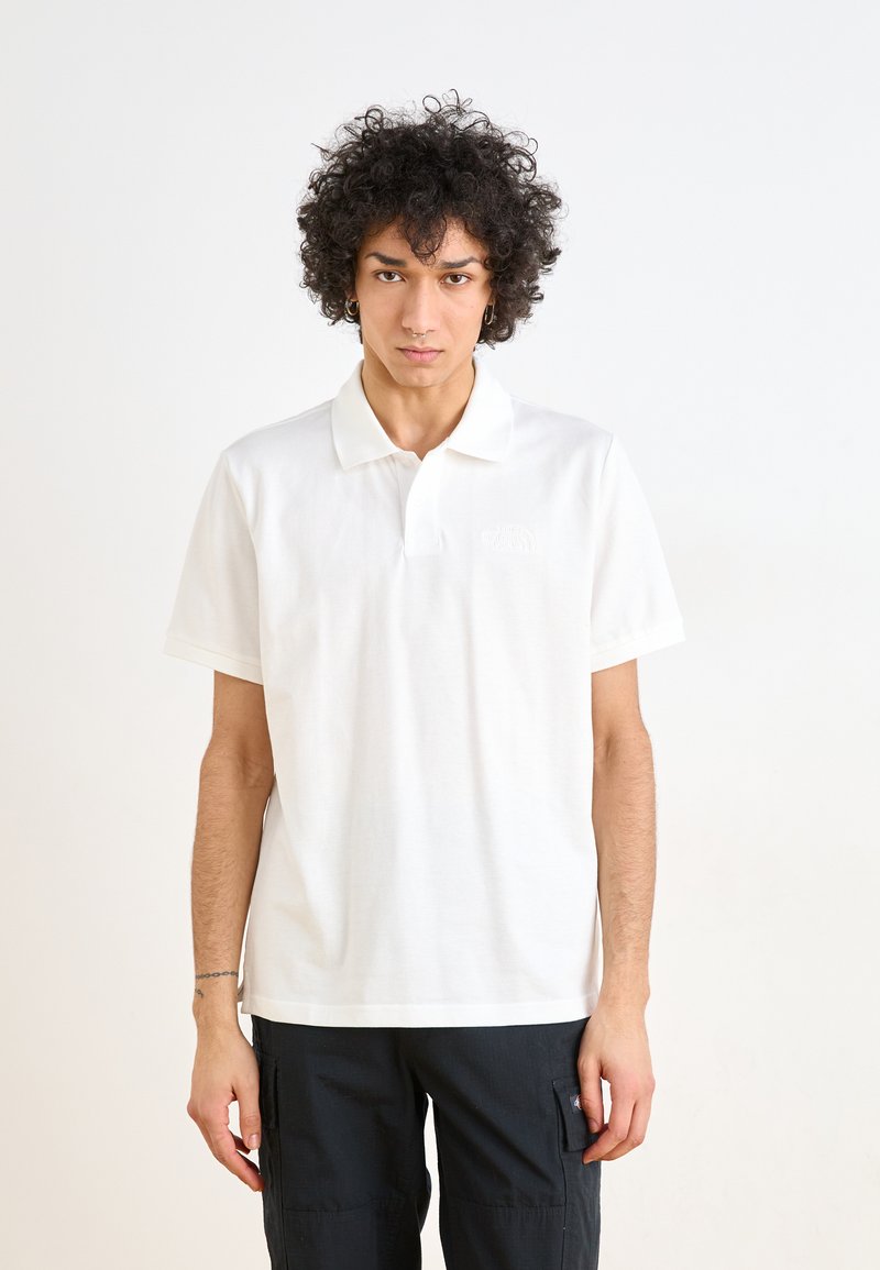 Polo-shirt blanc à manches courtes en coton, avec un col classique et un logo brodé discret sur la poitrine gauche. Pantalons cargo noirs portés.