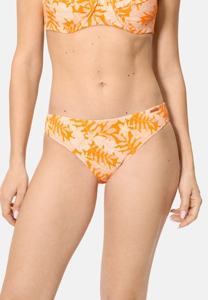 Mujer vistiendo bikini de flores naranja y beige, mostrando la zona media y las manos contra un fondo neutro.