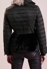 Osoba w czarnej pikowanej kurtce z panelami z futra sztucznego i paskiem, w połączeniu z czarnymi skórzanymi spodniami, pokazana od tyłu.