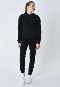 Zwarte hoodie met trekkoorden en een voorzak, gecombineerd met bijpassende zwarte joggers met elastische boorden, aangevuld met witte sneakers.