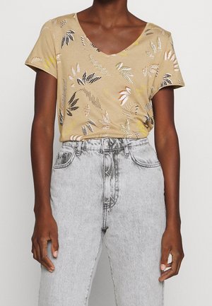 Vrouw draagt een beige T-shirt met bloemenprint en V-hals, ingestopt in een hoogwater lichtegrijze spijkerbroek.