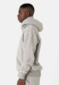 Grijze hoodie gemaakt van zacht materiaal, met witte decoratieve stiksels langs de naden en een kangoeroezak aan de voorkant.