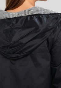 Černá větrovka s texturovaným stříbrným síťovaným límcem. Má kapuci a hladký, lesklý vnější povrch.