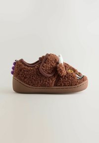 Chaussure pour enfant marron en peluche avec un visage de monstre, détail des cornes blanches, pointes violettes à l'arrière et fermeture à bande Velcro sur un fond neutre.
