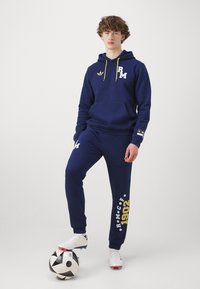 Conjunto de hoodie azul marinho e calças de treino feitas de tecido texturizado, com cordões amarelos, logos e detalhes em texto branco. Bola de futebol preta e branca.