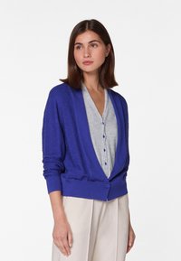 Paarse cardigan met een relaxte pasvorm, lange mouwen en een enkele knoopsluiting, gelaagd over een lichtgrijs topje met knopen. Zachte textuur.