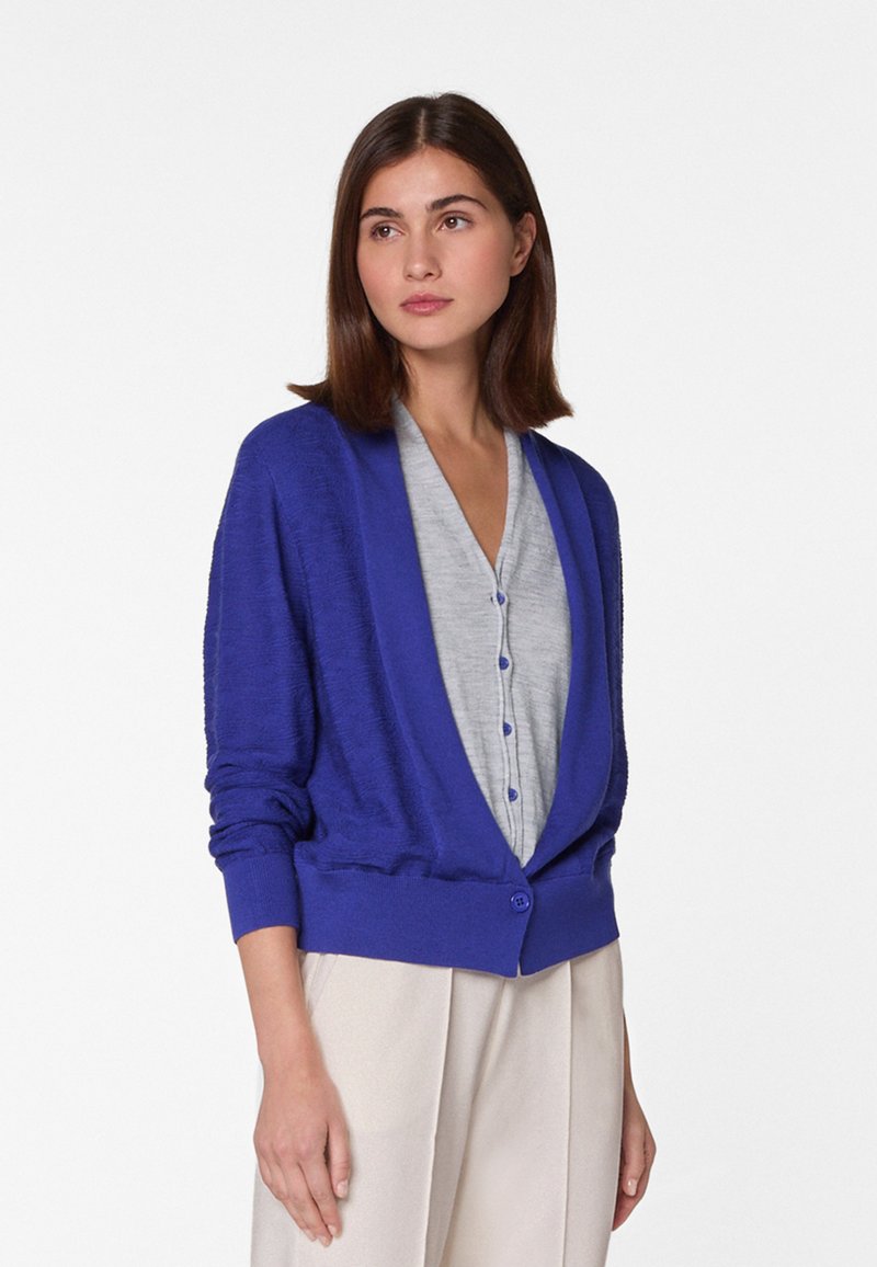 Paarse cardigan met een relaxte pasvorm, lange mouwen en een enkele knoopsluiting, gelaagd over een lichtgrijs topje met knopen. Zachte textuur.