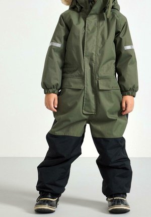 Combinaison de neige imperméable verte avec capuche, bandes réfléchissantes sur les manches, poches avant, et jambes inférieures noires ; tissu texturé, chaussures d'hiver.