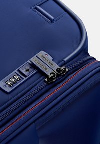 Valigia rigida blu navy con lucchetto a combinazione, doppia cerniera con dettagli rossi e superficie testurizzata per una maggiore durata.