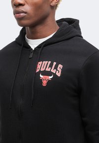 Μαύρο φούτερ με φερμουάρ, κόκκινο κείμενο "BULLS" και λογότυπο κεφαλής ταύρου στο αριστερό στήθος, φορεμένο πάνω από λευκό μπλουζάκι με στρογγυλή λαιμόκοψη.