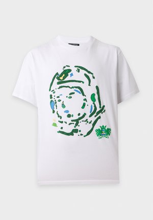 Homme debout, les mains dans les poches, portant un T-shirt blanc avec un imprimé abstrait de visage vert, pantalon noir et baskets noir et blanc.