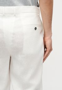 Homme portant un pantalon en lin blanc avec une poche arrière boutonnée, debout avec le bras droit détendu le long du corps.