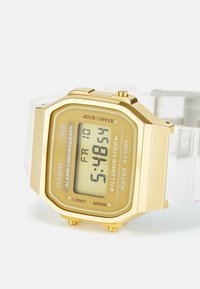 Montre numérique avec un boîtier en acier inoxydable doré, un bracelet transparent, un affichage LED, et des fonctionnalités telles que l'alarme, le chronomètre et la résistance à l'eau.