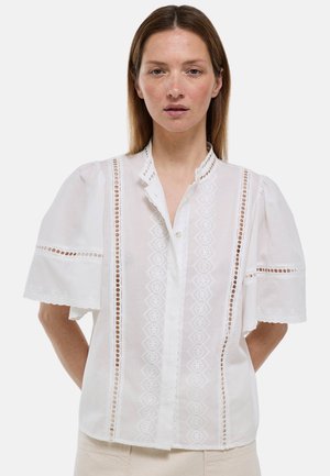 Femme portant un chemisier blanc à manches courtes avec des panneaux en dentelle et des bords festonnés, debout les mains derrière le dos sur un fond uni.