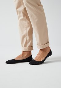 Ballerines noires en tissu doux, avec un design décolleté et un motif texturé sur la zone des orteils, associées à un pantalon beige clair.