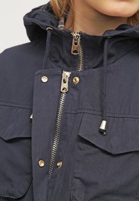 Navy blue parka jacket with a gold zipper, snap buttons, and drawstring hood. Features dual front pockets and a soft, textured fabric.